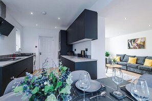 Une cuisine moderne aux meubles sombres, un coin repas avec une table dressée pour quatre personnes, et un salon avec un canapé et un tableau accroché au mur.