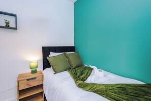 A single bed with a green blanket, a bedside table with a lamp, and a wall-mounted picture frame. - M22 | Near Airport | Parking | WiFi | 15% Off (Wythenshawe)