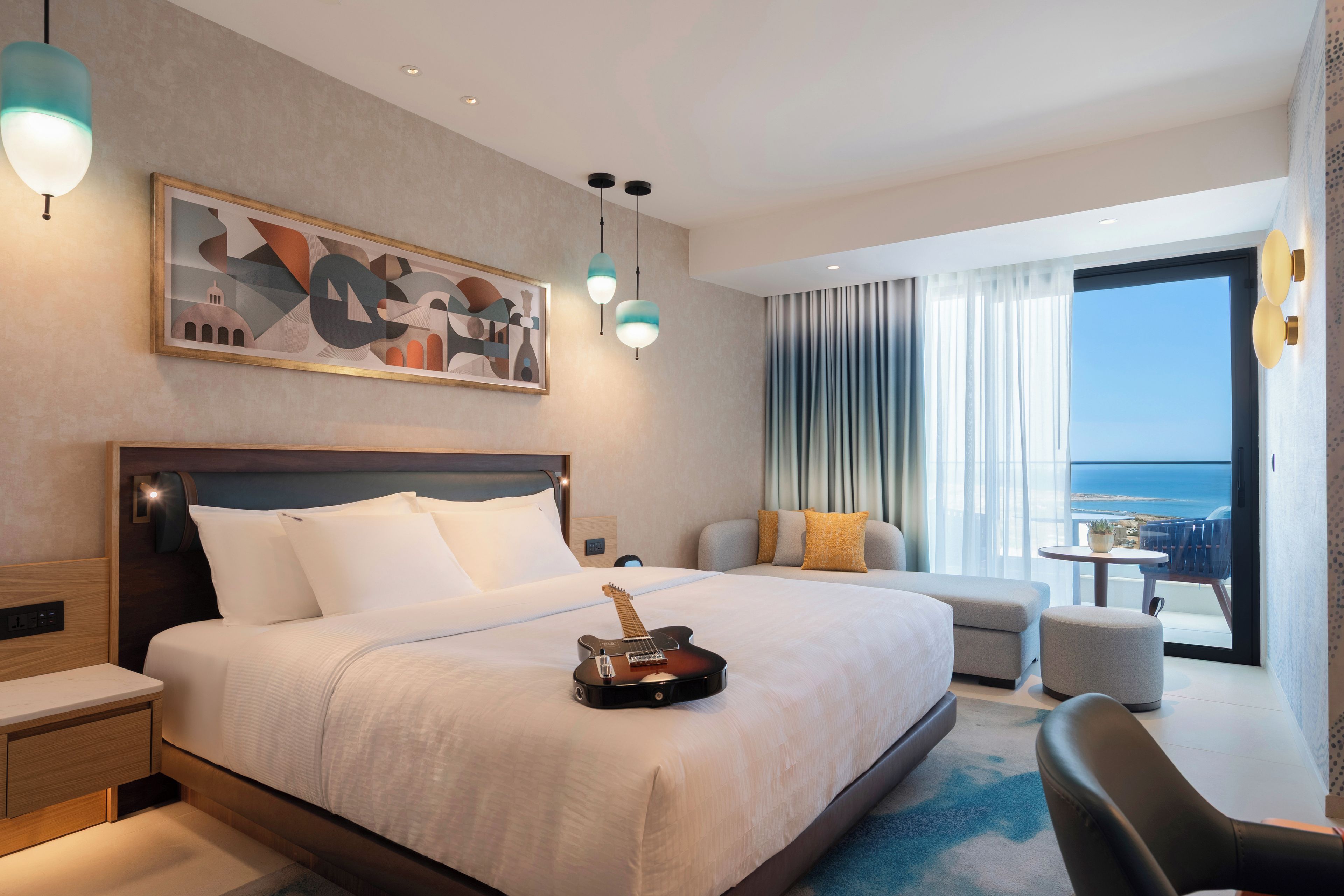 A hotel room with a large bed, a painting, a guitar, a sofa, and a view of the sea.