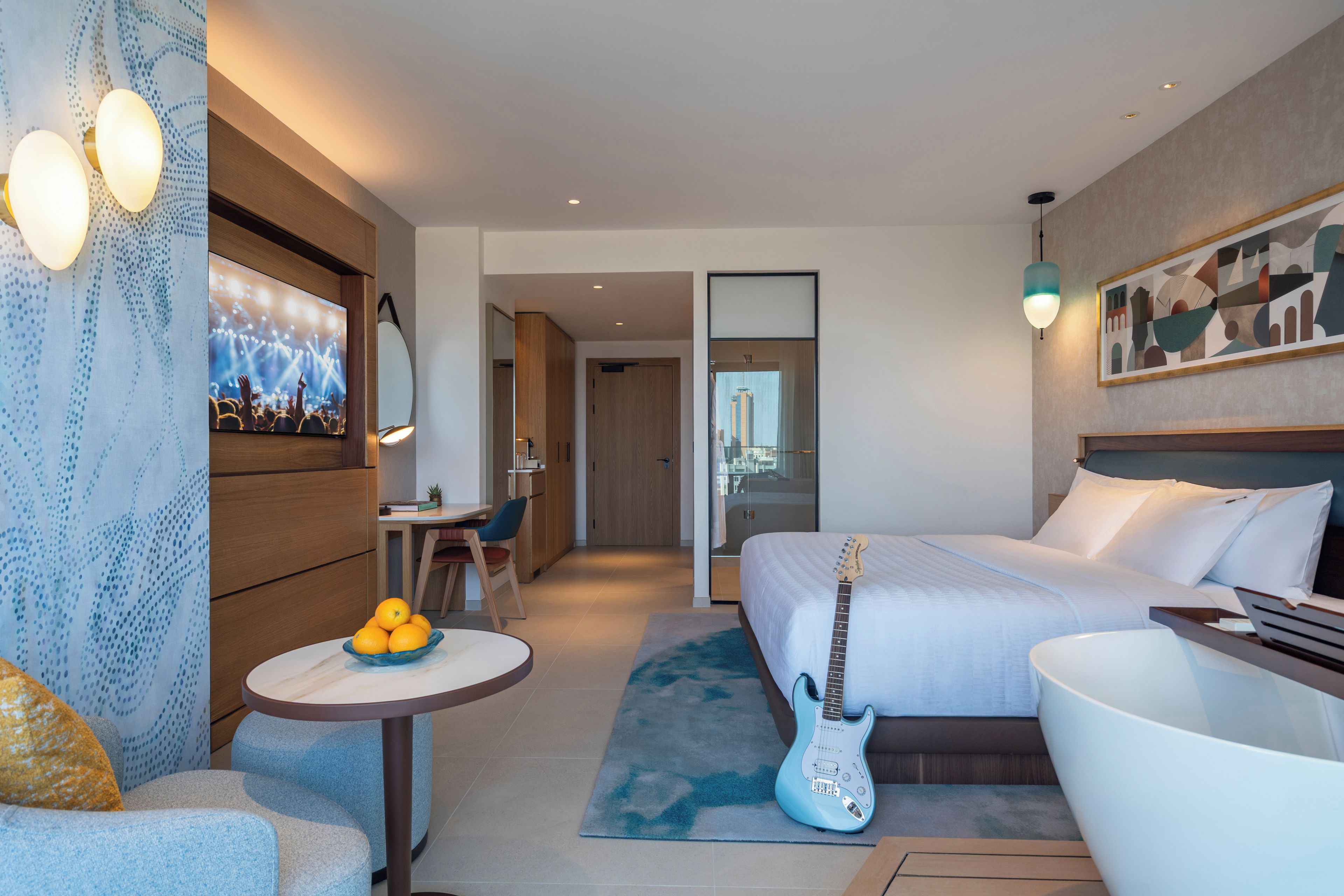 A modern hotel room with a bed, a desk, a chair, a guitar, and a television on the wall.