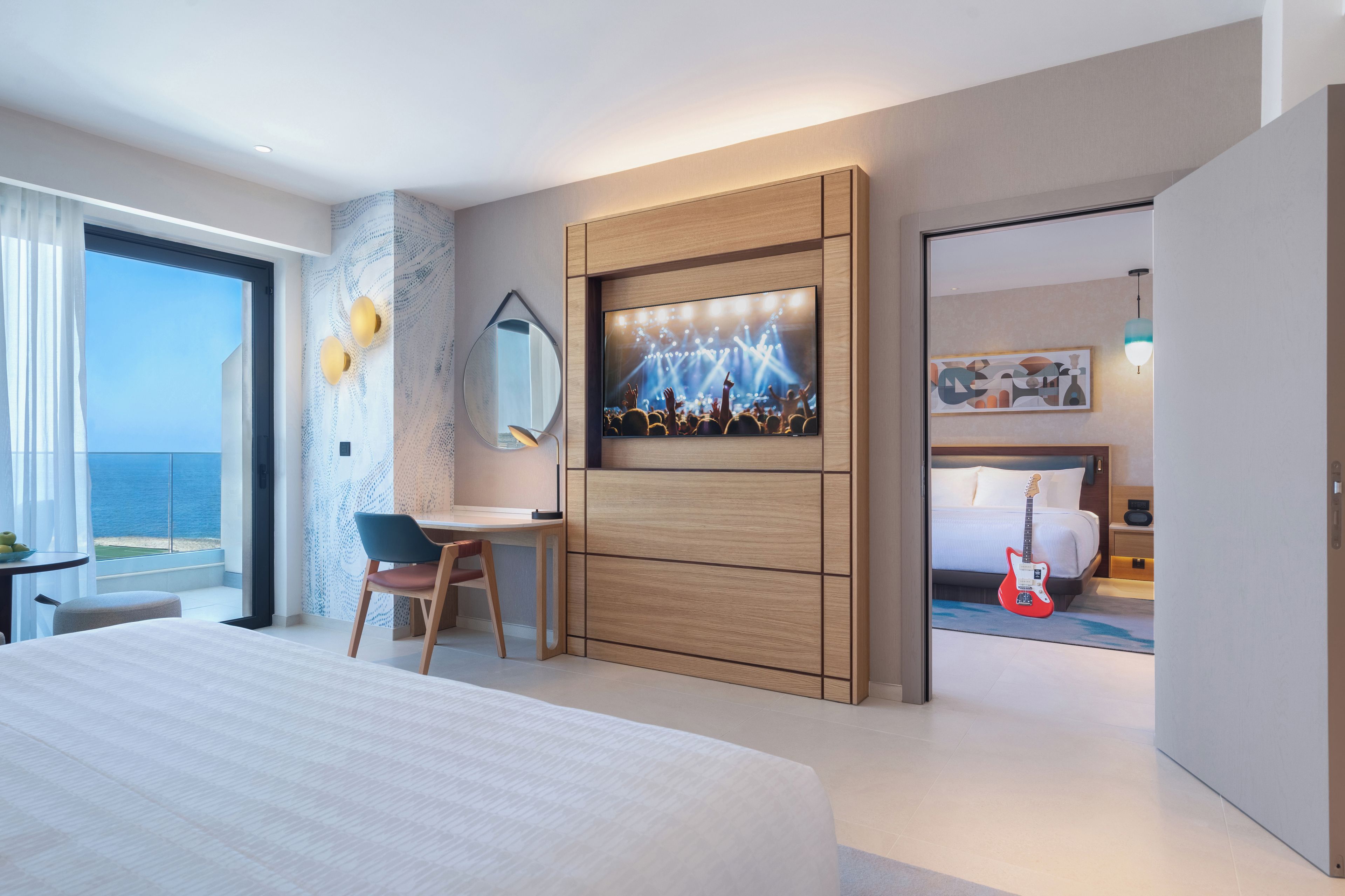 A modern hotel room with a large bed, a desk, a chair, a TV, and a view of the sea.