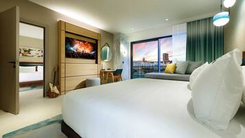 A hotel room with a large bed, a desk, a chair, a guitar, a painting, and a view of the city.