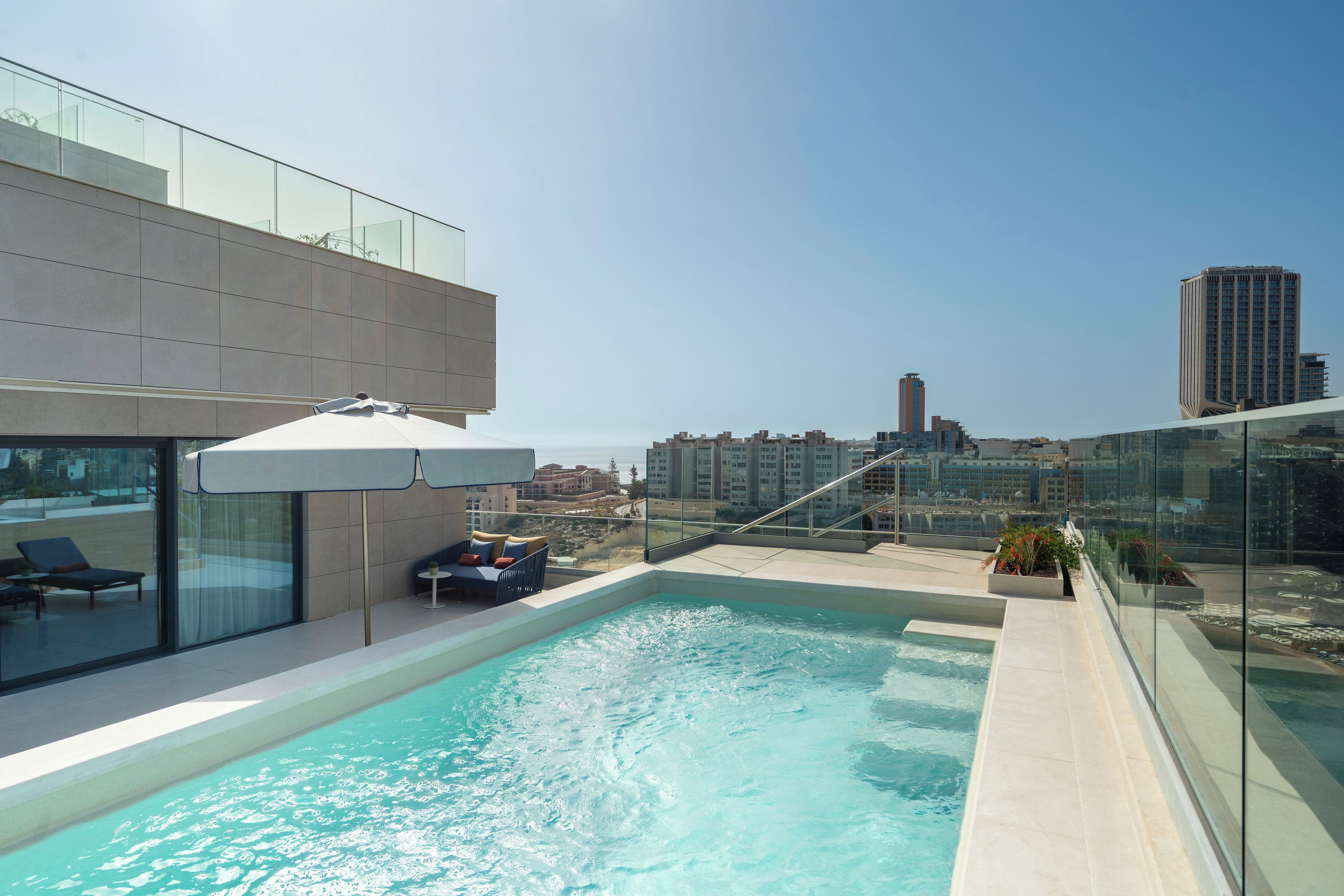 A rooftop pool with glass railings, a seating area with a table and chairs, and a view of the cityscape.