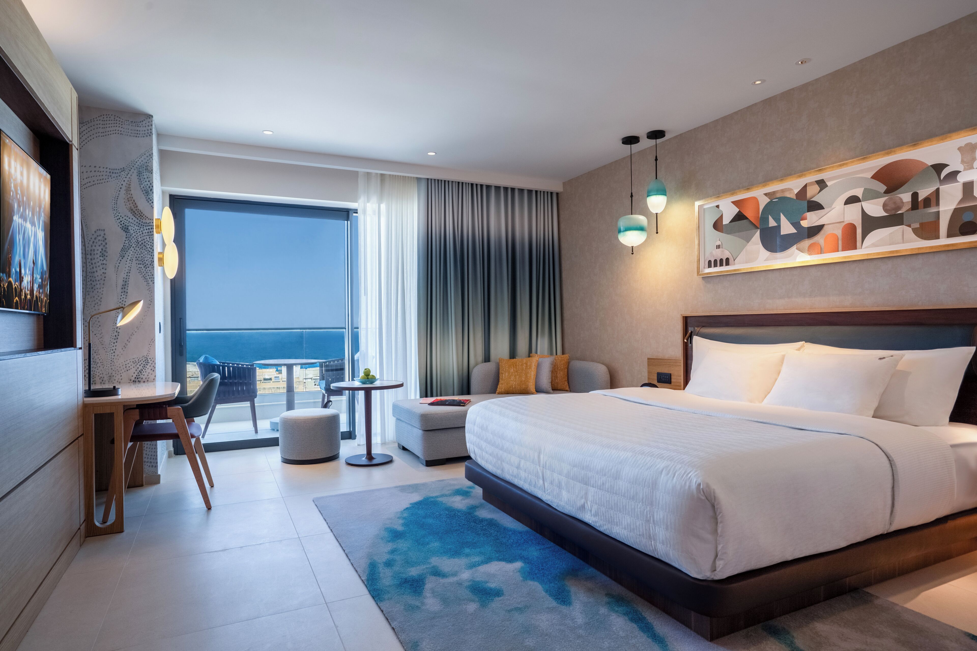 A modern hotel room with a large bed, a desk, a chair, a dining table, and a view of the sea.