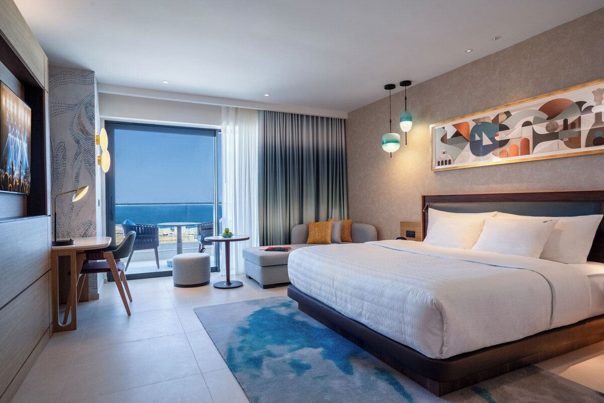 A modern hotel room with a large bed, a desk, a chair, a dining table, and a view of the sea.