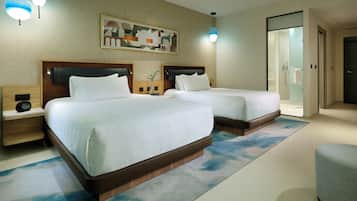 A hotel room with two beds, a nightstand with a speaker, a wall-mounted artwork, and a bathroom visible through a glass door.