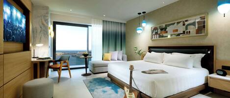 A modern hotel room with a large bed, a desk, a chair, a guitar, and a view of the city.
