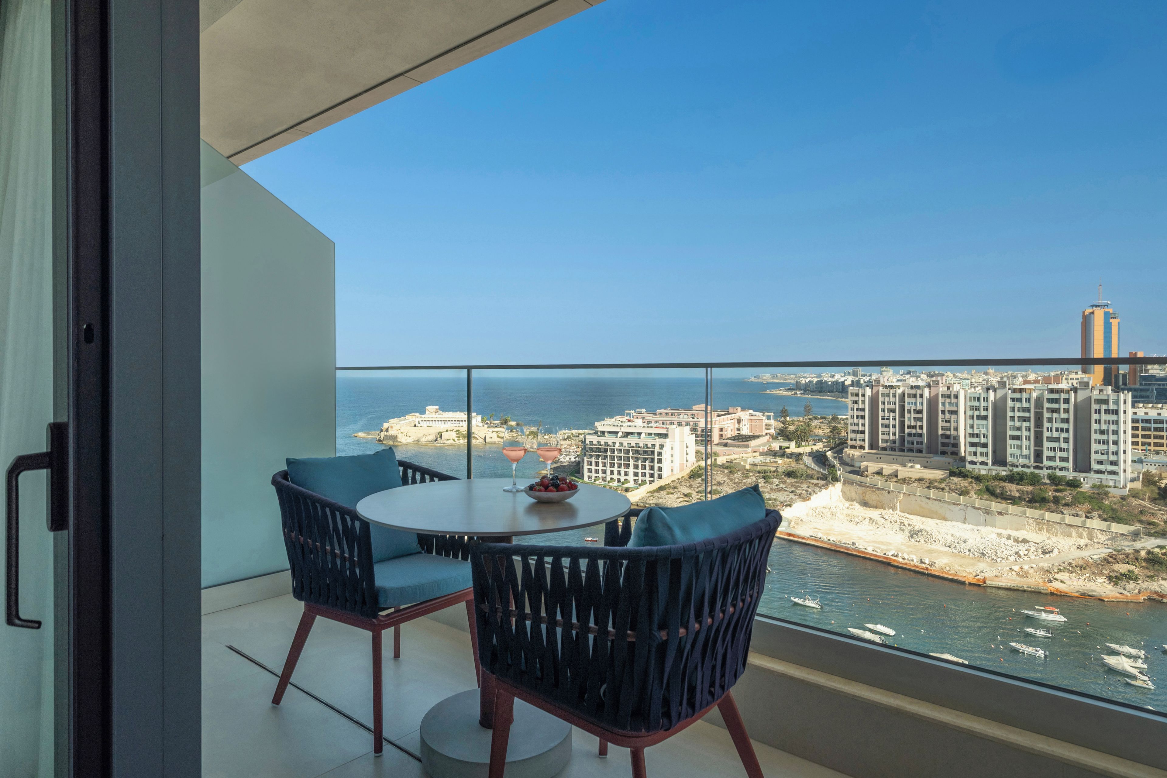 Uma varanda com mesa e cadeiras, com vista para um horizonte costeiro com edifícios e céu claro.
