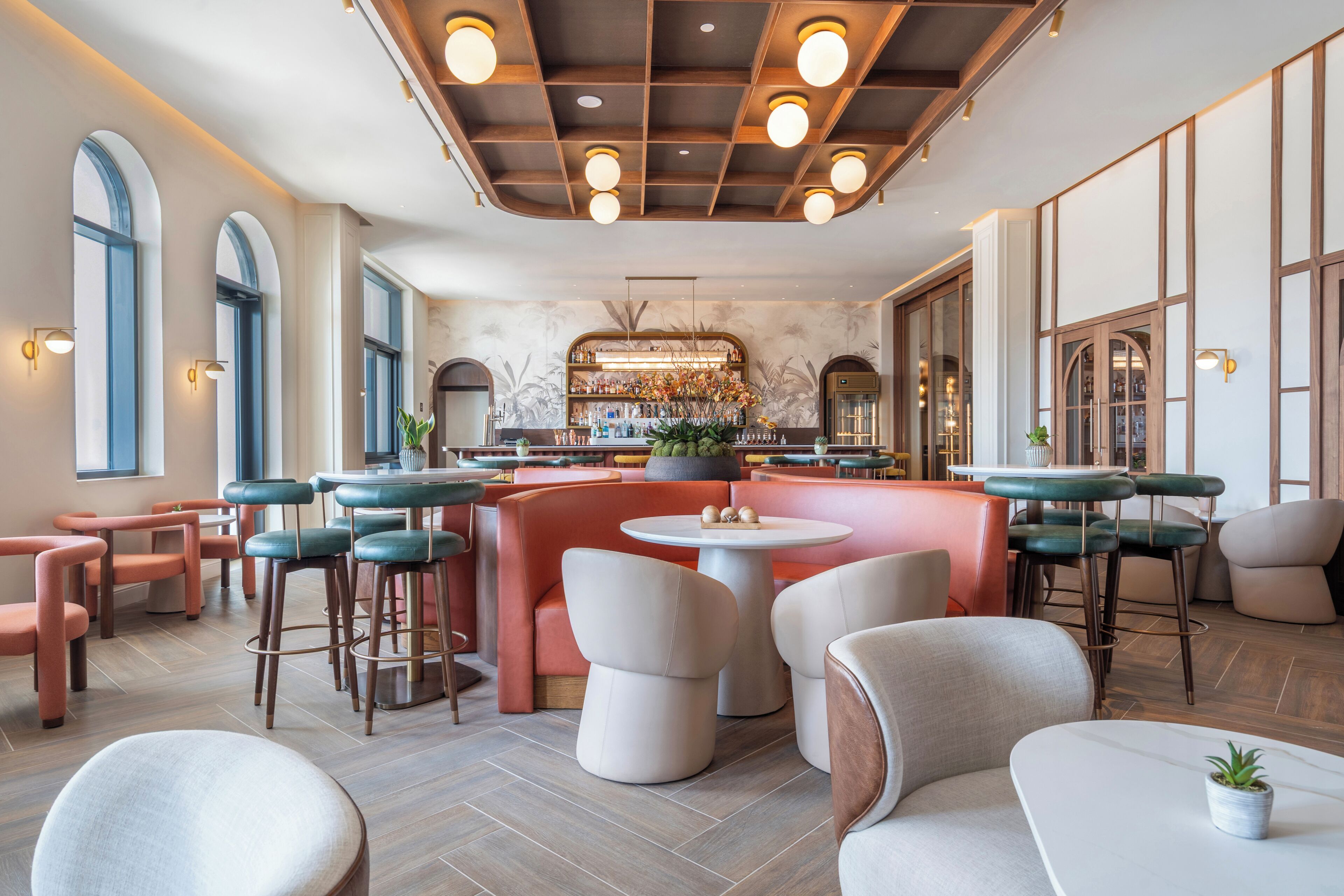 A modern bar area with round tables and bar stools, a central bar counter with a decorative plant, and a patterned wall in the background.