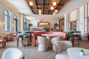 A modern bar area with round tables and bar stools, a central bar counter with a decorative plant, and a patterned wall in the background. - Hard Rock Hotel Malta (St. Julian's)