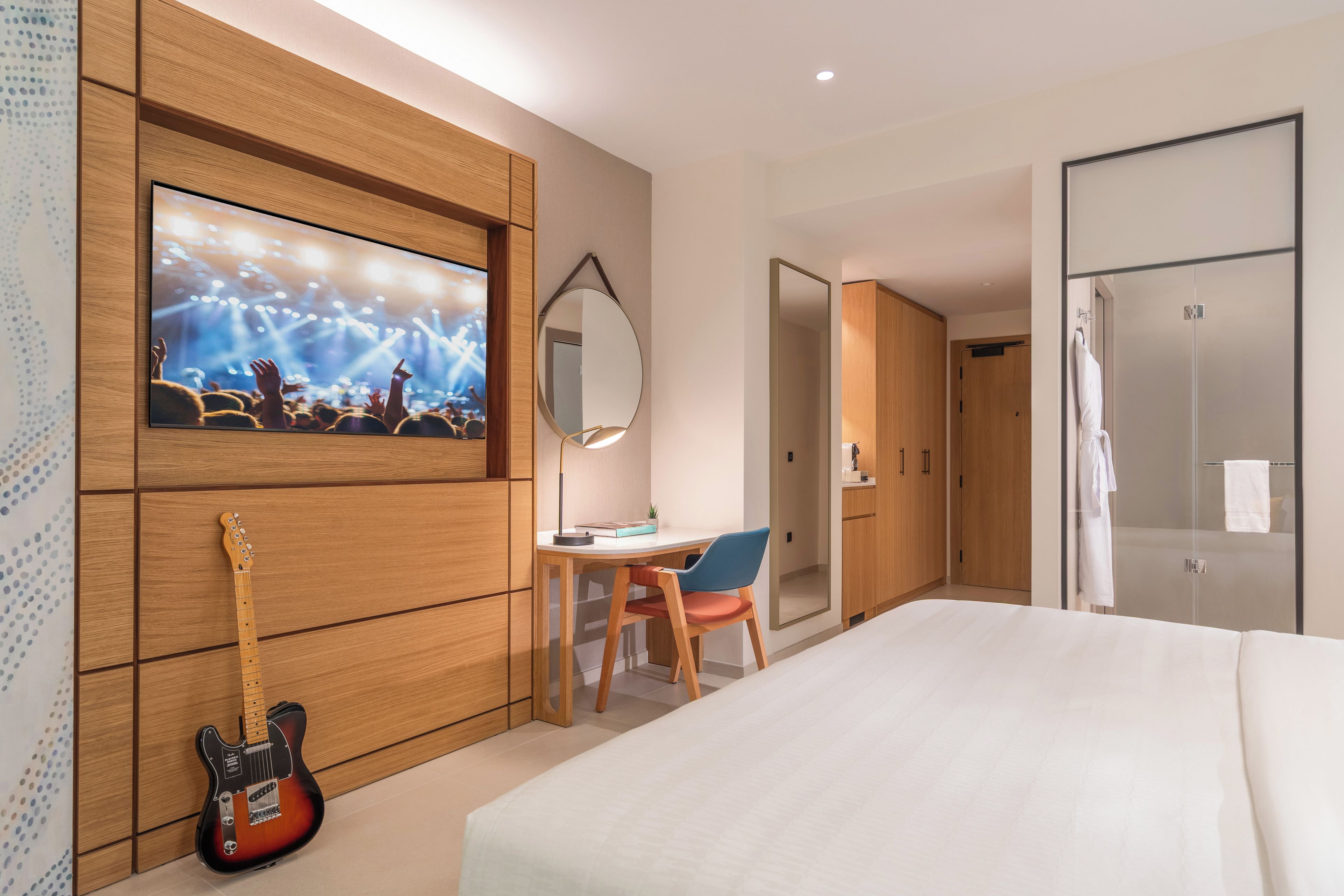 Een hotelkamer met een bed, een bureau met stoel, een gitaar, een aan de muur bevestigde televisie en een kledingkast.
