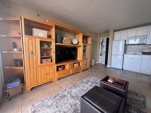 A living room with a wooden entertainment center, a flat-screen TV, and a kitchen area with white cabinets and appliances. - Family Fun * On the Ocean (Daytona Beach)