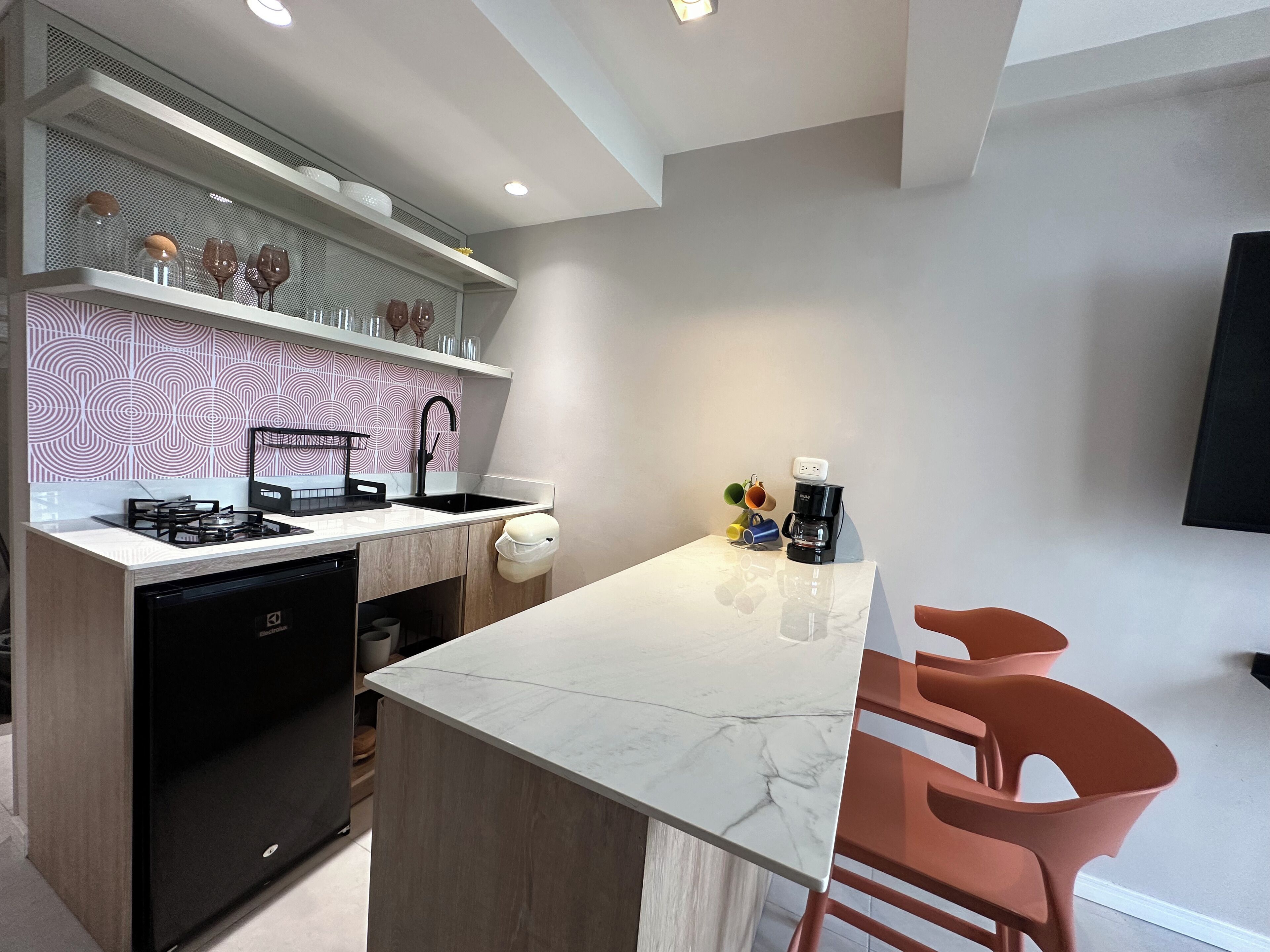 A modern kitchen with a built-in dishwasher, a gas stove, a sink, and a marble countertop. There are two orange chairs and a coffee maker on the countertop.