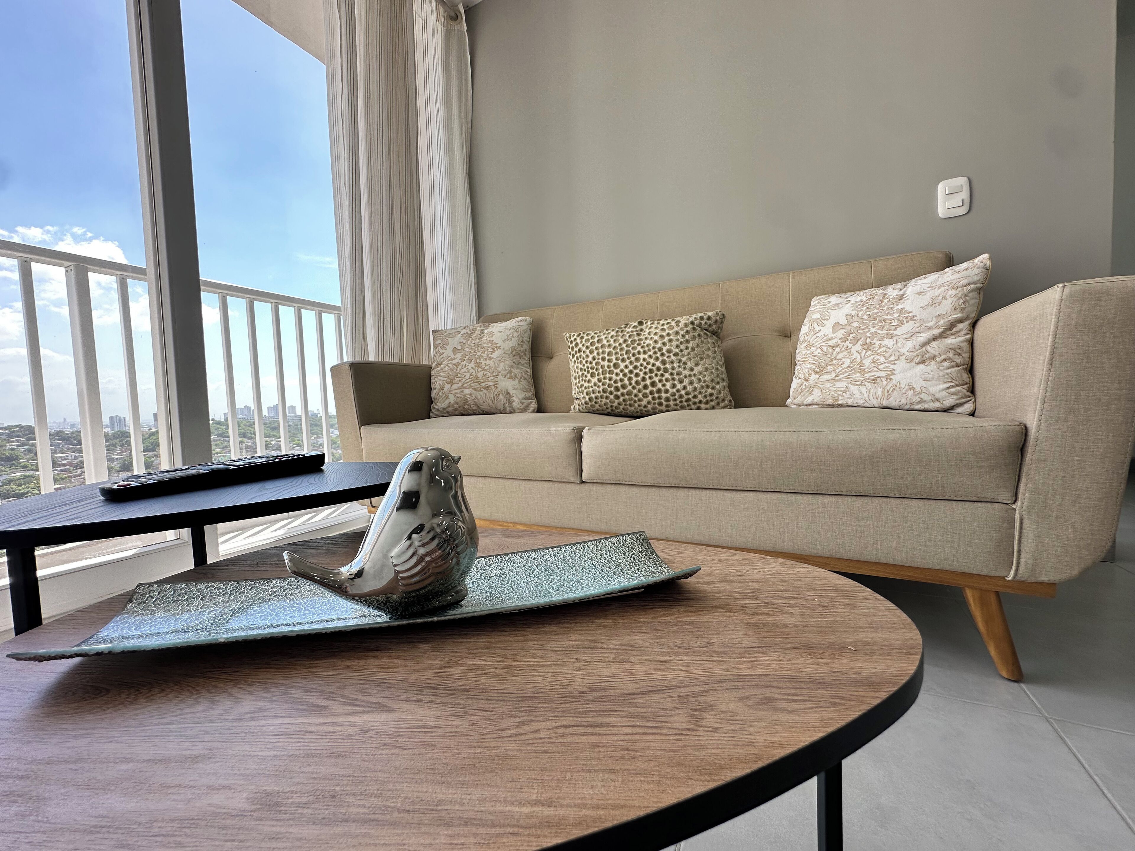 A living room with a beige sofa, patterned cushions, a remote control, and a decorative horse figurine on a round table.