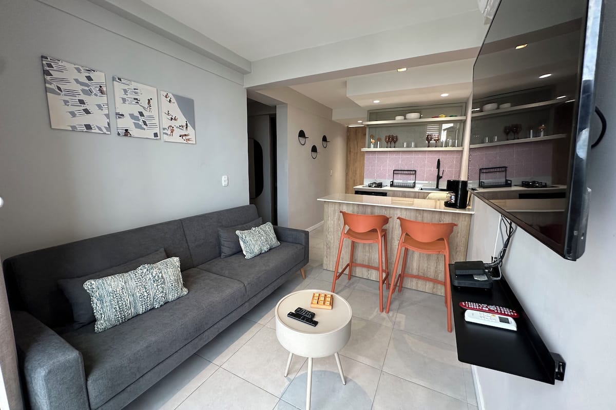 Uma sala de estar moderna com um sofá cinzento, uma mesa de centro e uma área de cozinha com balcão e dois bancos.