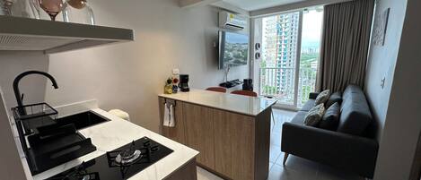 A modern kitchen with a gas stove, a coffee machine, and a small dining area. There is a sofa and a TV in the living area, with a view of high-rise buildings.