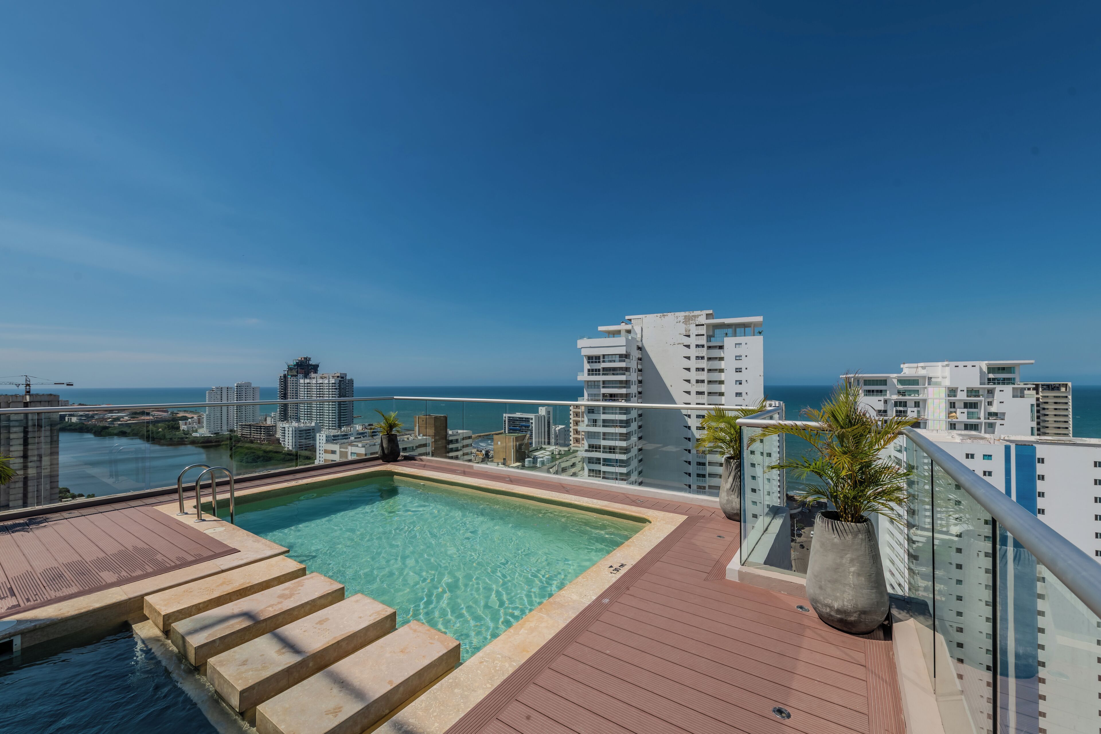 A rooftop pool with clear blue water, surrounded by a wooden deck and glass railings, overlooking a cityscape and the ocean.