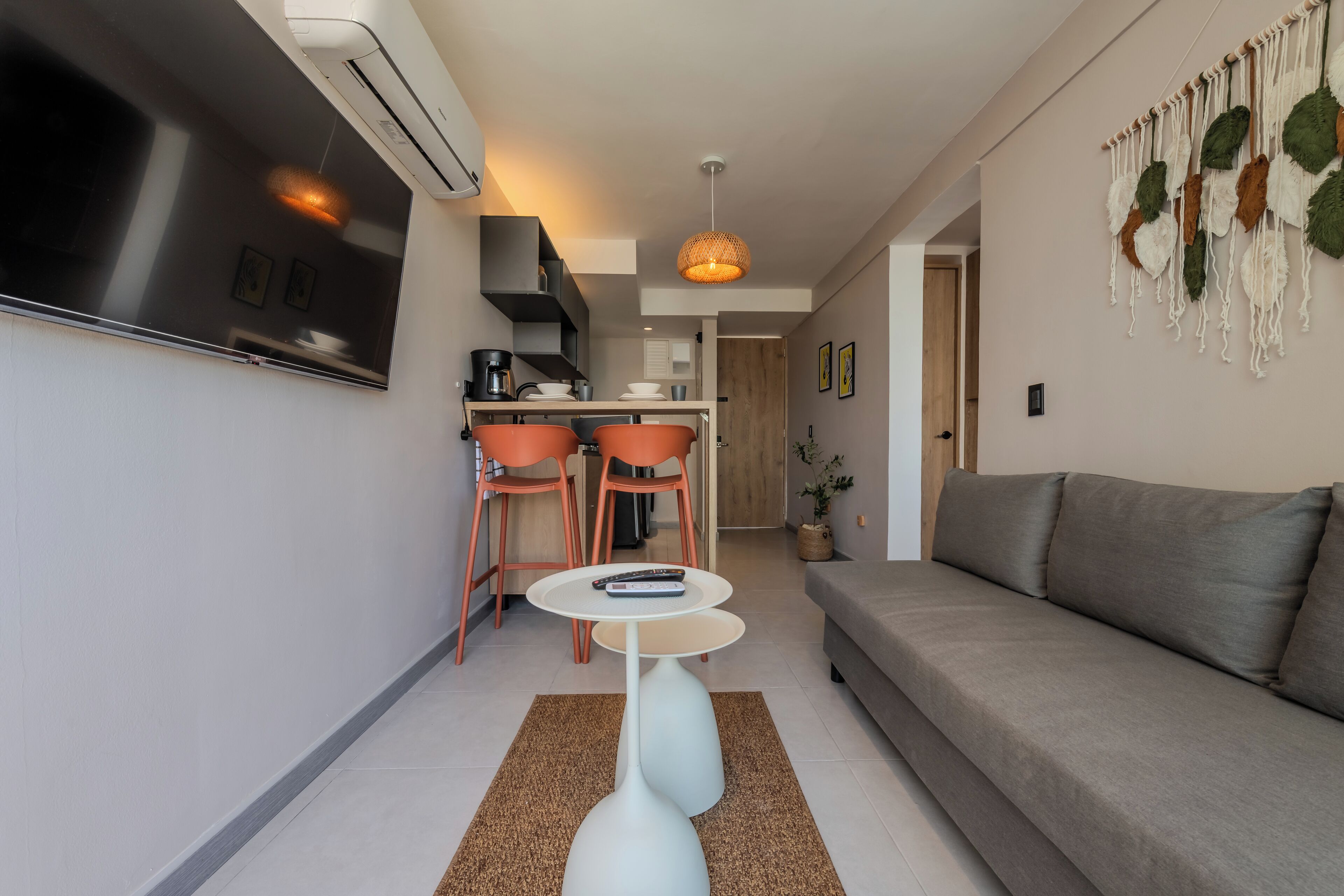 A modern living room with a grey sofa, a white coffee table, and orange chairs. There is a kitchen area with a black stove and a hanging light fixture.
