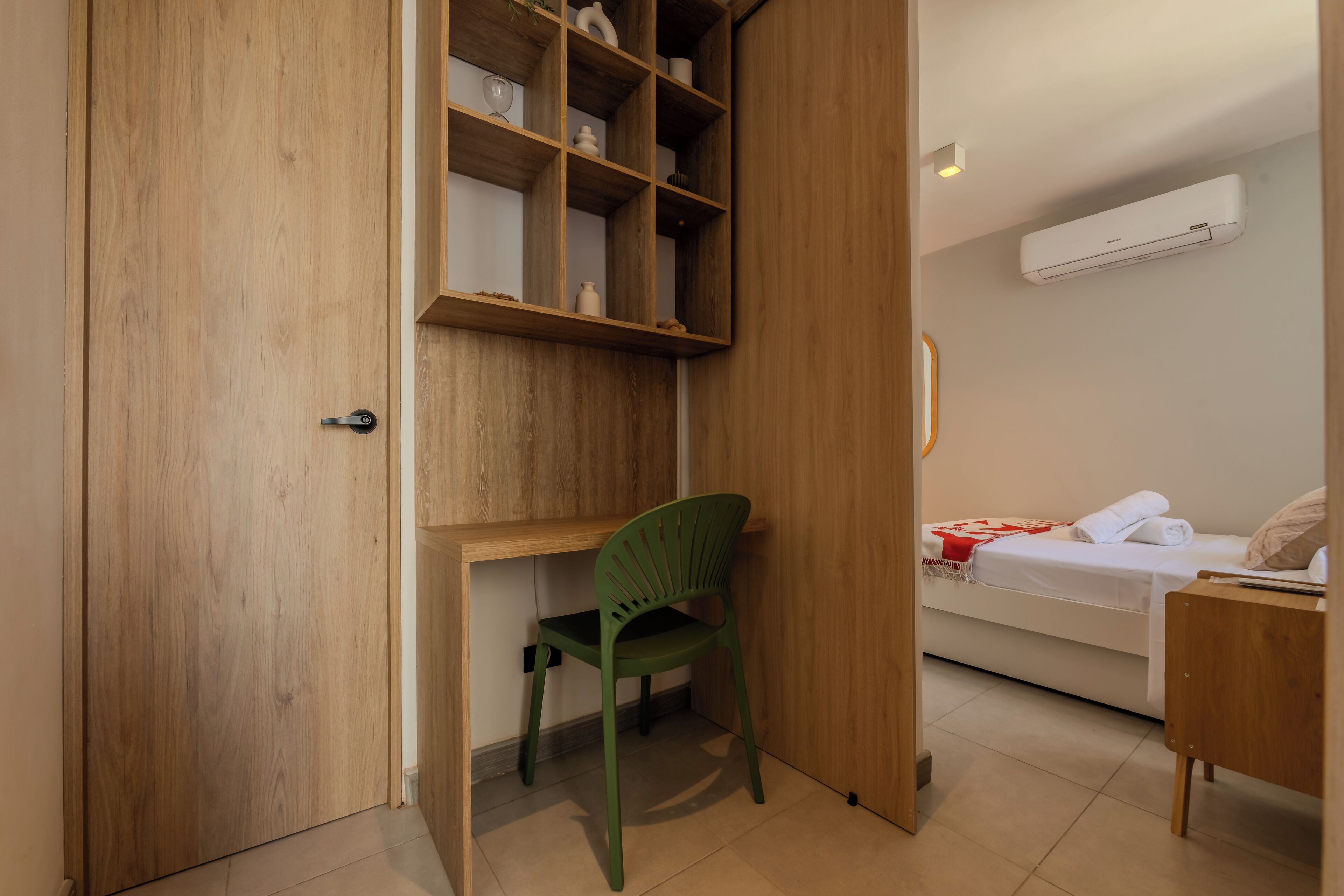 A room with a wooden door, a green chair, a bed with a white mattress and red blanket, and a white air conditioner on the wall.