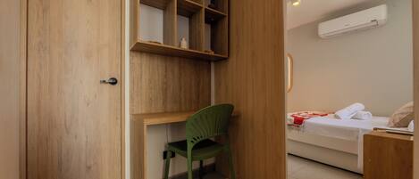 A room with a wooden door, a green chair, a bed with a white mattress and red blanket, and a white air conditioner on the wall.