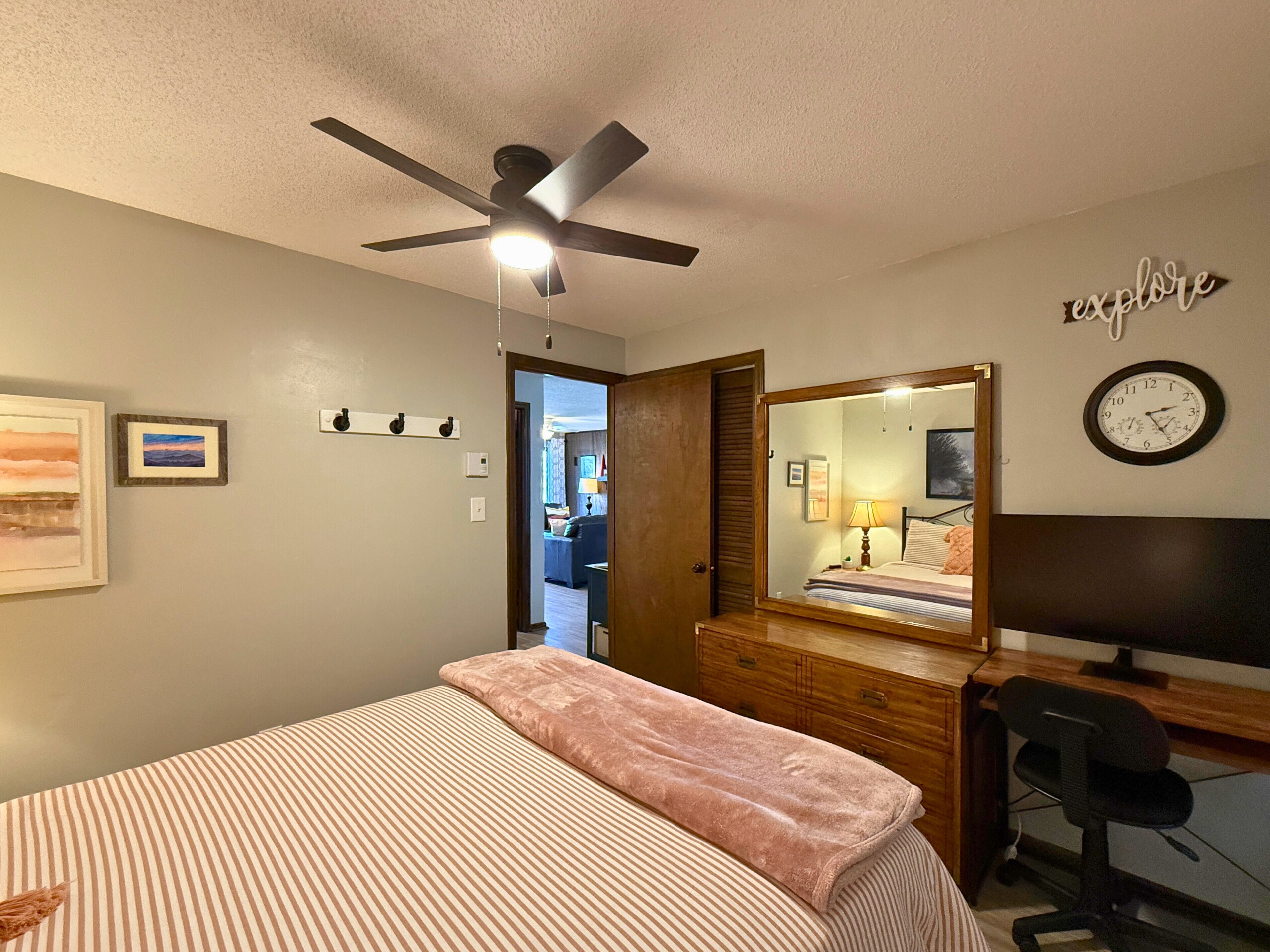 A bedroom with a bed, a desk, a chair, a ceiling fan, a mirror, and a clock on the wall.