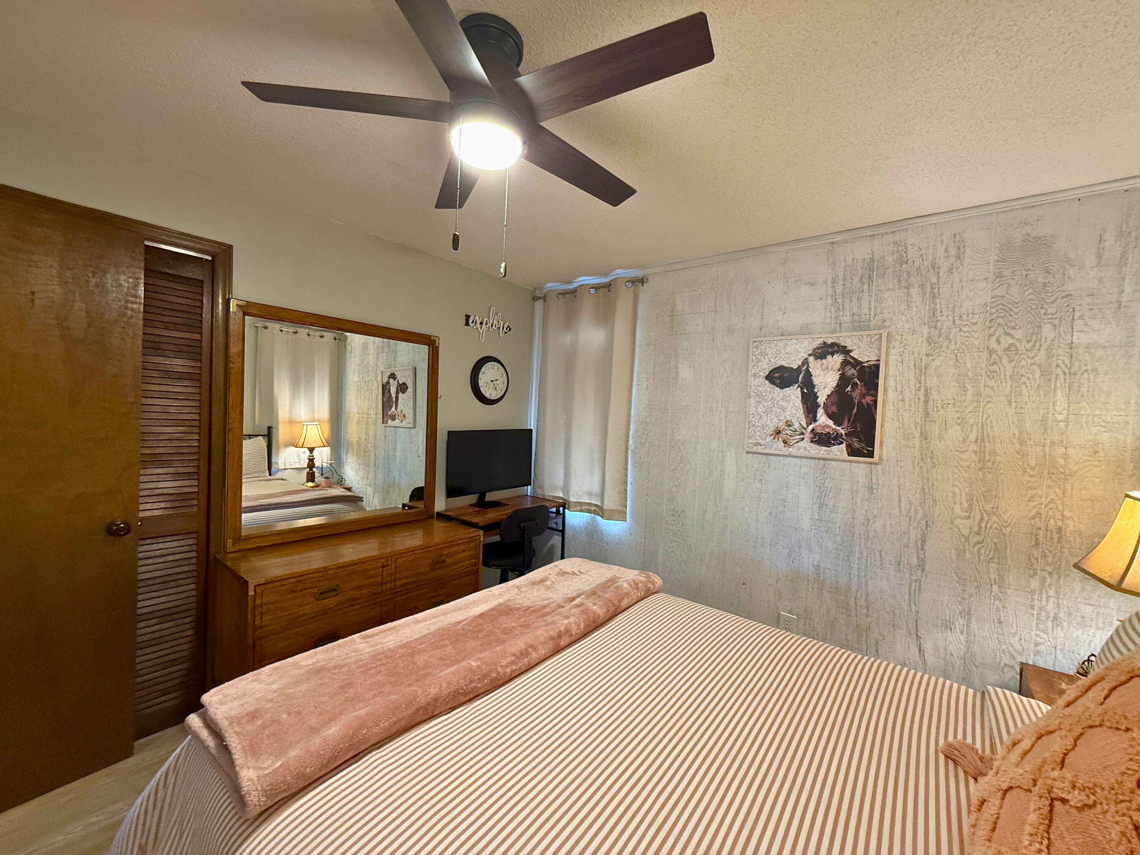 A bedroom with a bed, a desk, a computer, a ceiling fan, a mirror, a clock, and a cow painting on the wall.
