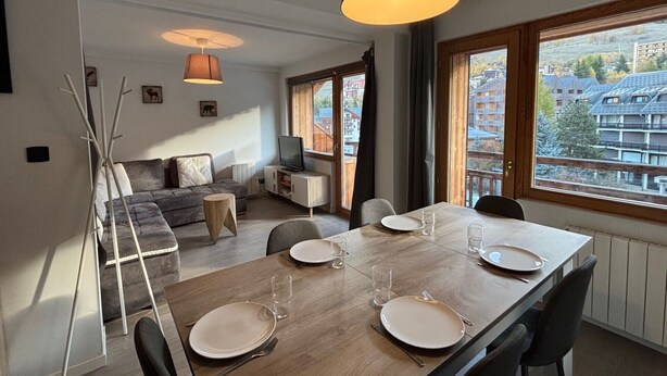A modern living room with a dining area, a sofa, a TV, and a large window offering a view of buildings. - Beautiful apartment, sleeps 5, mountain view (Les Deux Alpes)