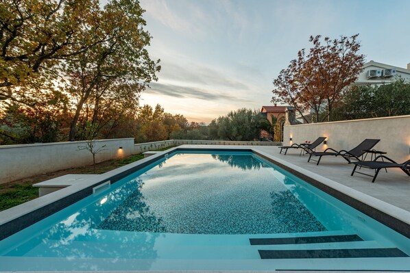 Ein rechteckiger Pool mit klar blauem Wasser, umgeben von einer niedrigen weißen Mauer. Auf der rechten Seite befinden sich Liegestühle mit Kissen und ein kleiner Tisch.