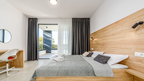 A modern hotel room with a large bed, a wooden headboard, a desk with a chair, and a view of the outdoors. - Villa The Family Zadarvillas (Jasenice)
