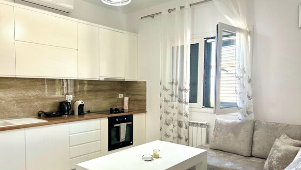 Una cocina moderna con armarios blancos, un horno negro y una cafetera. Hay una mesa blanca con una vela y una ventana con cortinas.