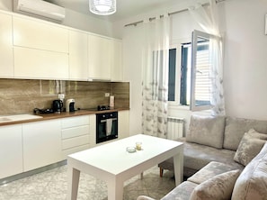 Una cocina moderna con armarios blancos, un horno negro y una cafetera. Hay una mesa blanca con una vela y una ventana con cortinas.