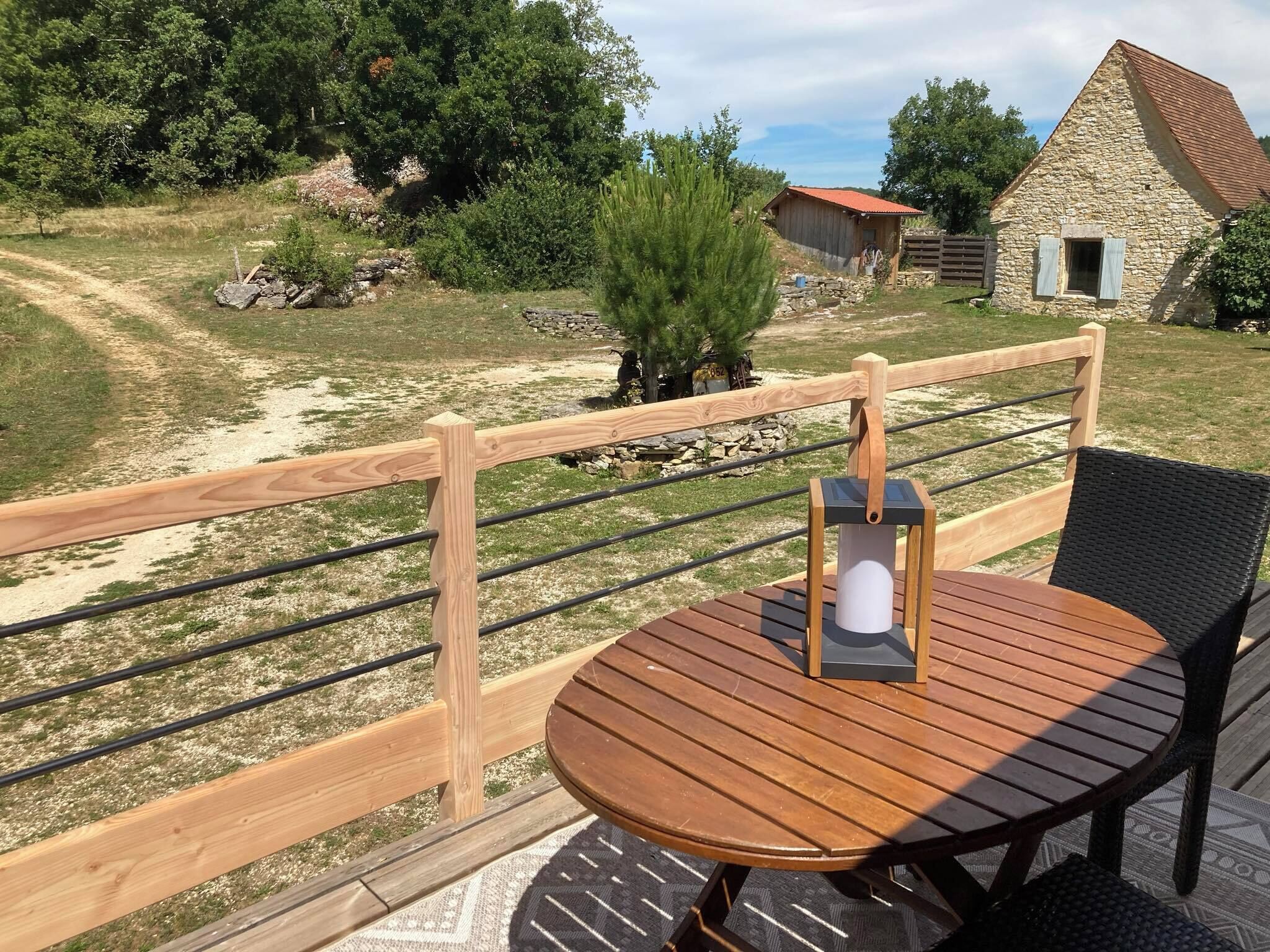 Une terrasse en bois avec une table ronde et une chaise, d’où l’on peut admirer un paysage rural avec un bâtiment en pierre et un chemin de terre.