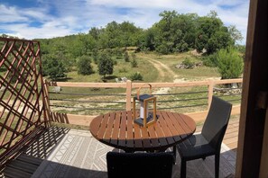 A wooden deck with a round table and a chair, overlooking a grassy area with trees and a dirt path. - Le PERCHOIR, comfortable gîte (Bouzic)