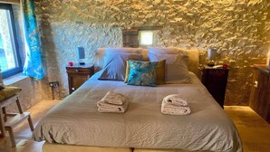 A bedroom with a stone wall, a bed with a grey bedspread, two towels on the bed, a nightstand with a lamp, and a window with curtains. - Le PERCHOIR, comfortable gîte (Bouzic)