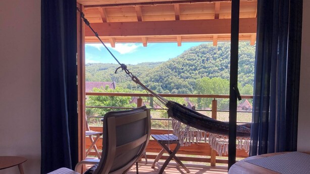 A balcony with a hammock, a cushioned chair, and a table with a view of a forested landscape. - Le PERCHOIR, comfortable gîte (Bouzic)
