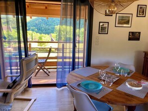 A dining area with a wooden table set for two, a view of a forested landscape through a glass door, and a ceiling fan. - Le PERCHOIR, comfortable gîte (Bouzic)