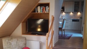 A cozy loft bedroom with a sloped ceiling, a television, a bookshelf, a dining area, and a kitchen in the background. - Apartment 4 with Balcony and Wi-Fi (Horumersiel)
