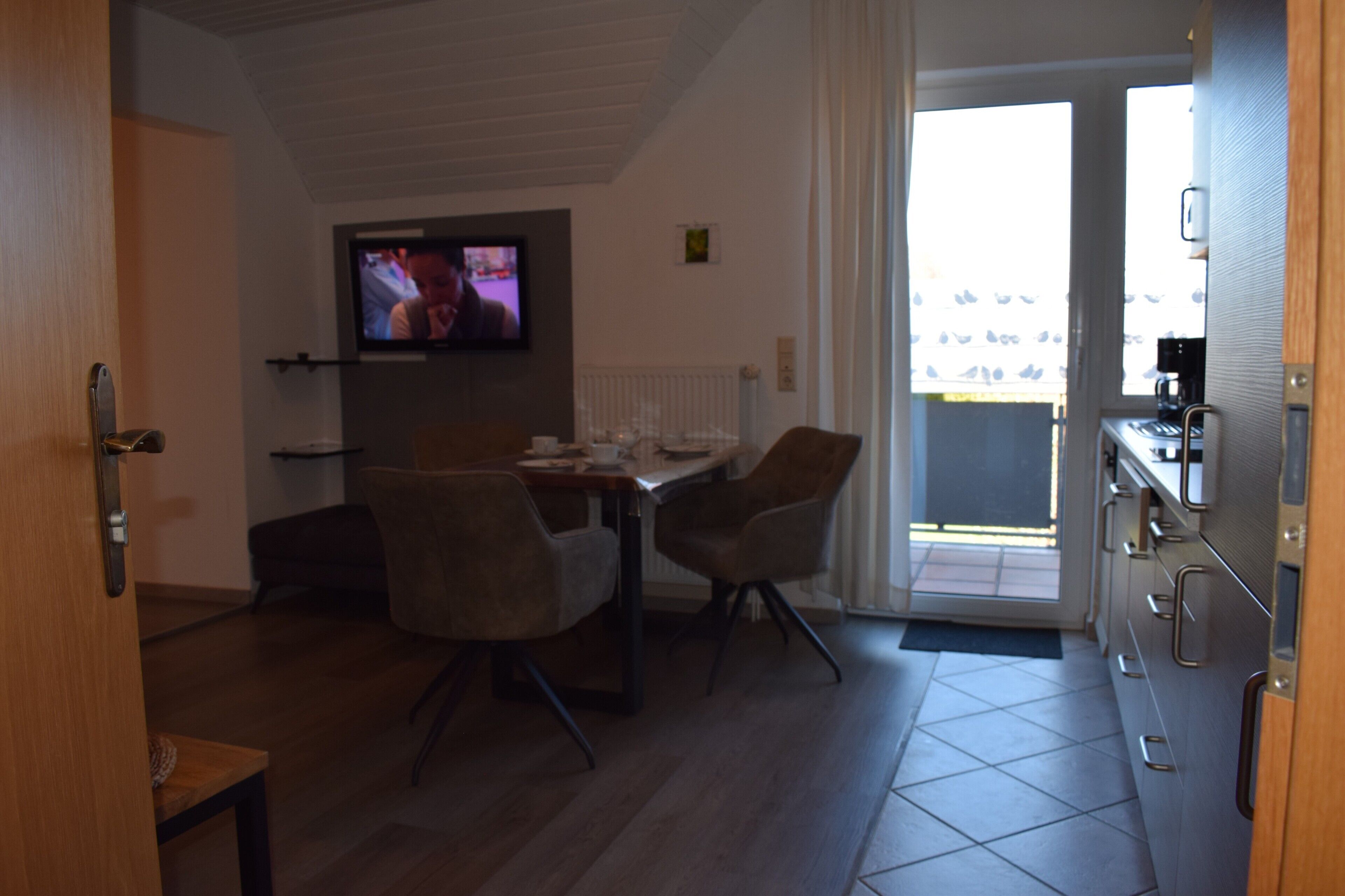 Zona pranzo con televisore, sedie, tavolo e angolo cottura con pensili e macchina per il caffè.