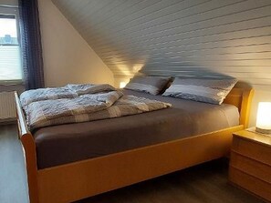 A bedroom with a sloped ceiling, a bed with a wooden frame, a bedside table with a lamp, and a window with curtains.