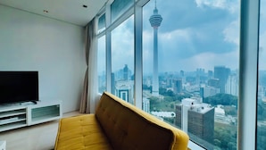 Una habitación con un sofá amarillo, un televisor sobre un soporte blanco y un ventanal grande con vista a la ciudad, donde destaca una torre.