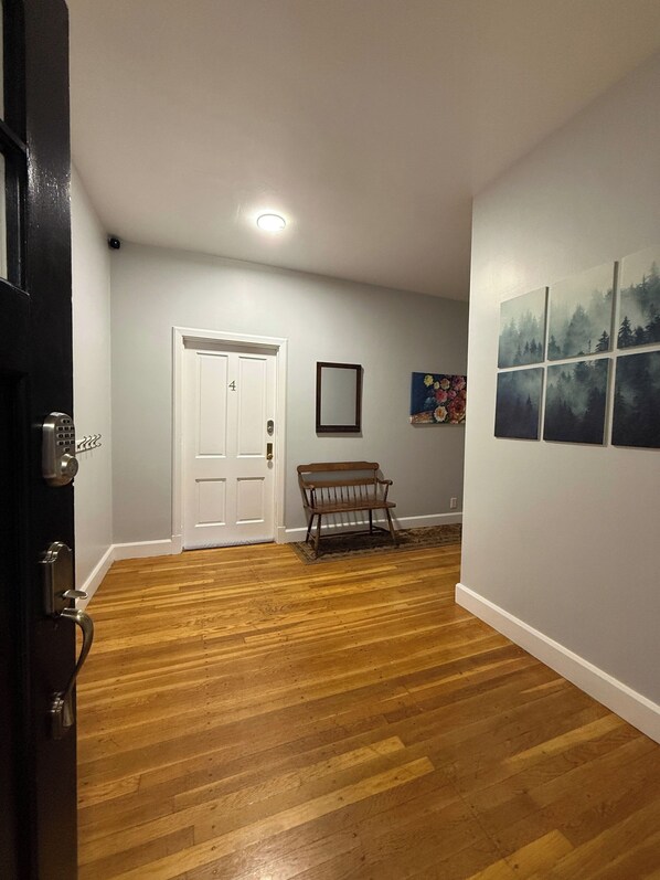 A hallway with wooden flooring, a white door, a bench, and a wall with a framed picture and a series of square paintings.