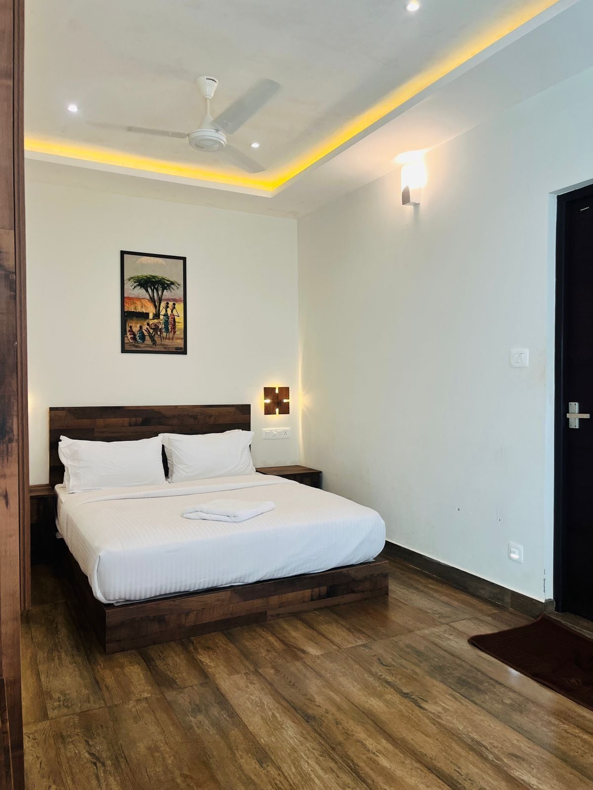 A bedroom with a wooden bed, a ceiling fan, a wall-mounted light, and a painting of a village scene.
