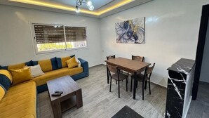 A modern living room with a sofa, coffee table, dining table, and chairs. There is a window with blinds, a ceiling light fixture, and a marble accent wall. - Bonne étoile Imsouane : ground floor Apartment (Imsouane,)