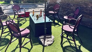 Eine Terrasse im Freien mit Tisch und Stühlen unter einem pinken Sonnenschirm, umgeben von Steinmauern und Grünpflanzen.
