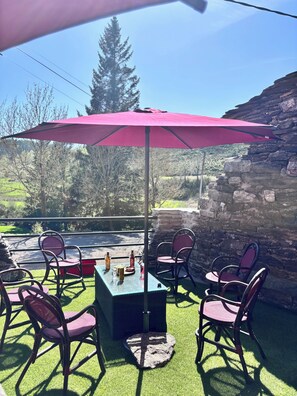 Eine Terrasse im Freien mit Tisch und Stühlen unter einem pinken Sonnenschirm, umgeben von Steinmauern und Grünpflanzen.