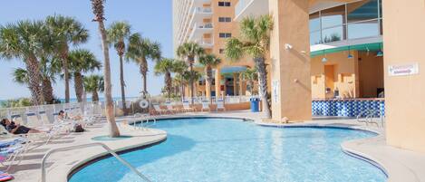 A pool area with lounge chairs, palm trees, and a building with a sign indicating a restroom.