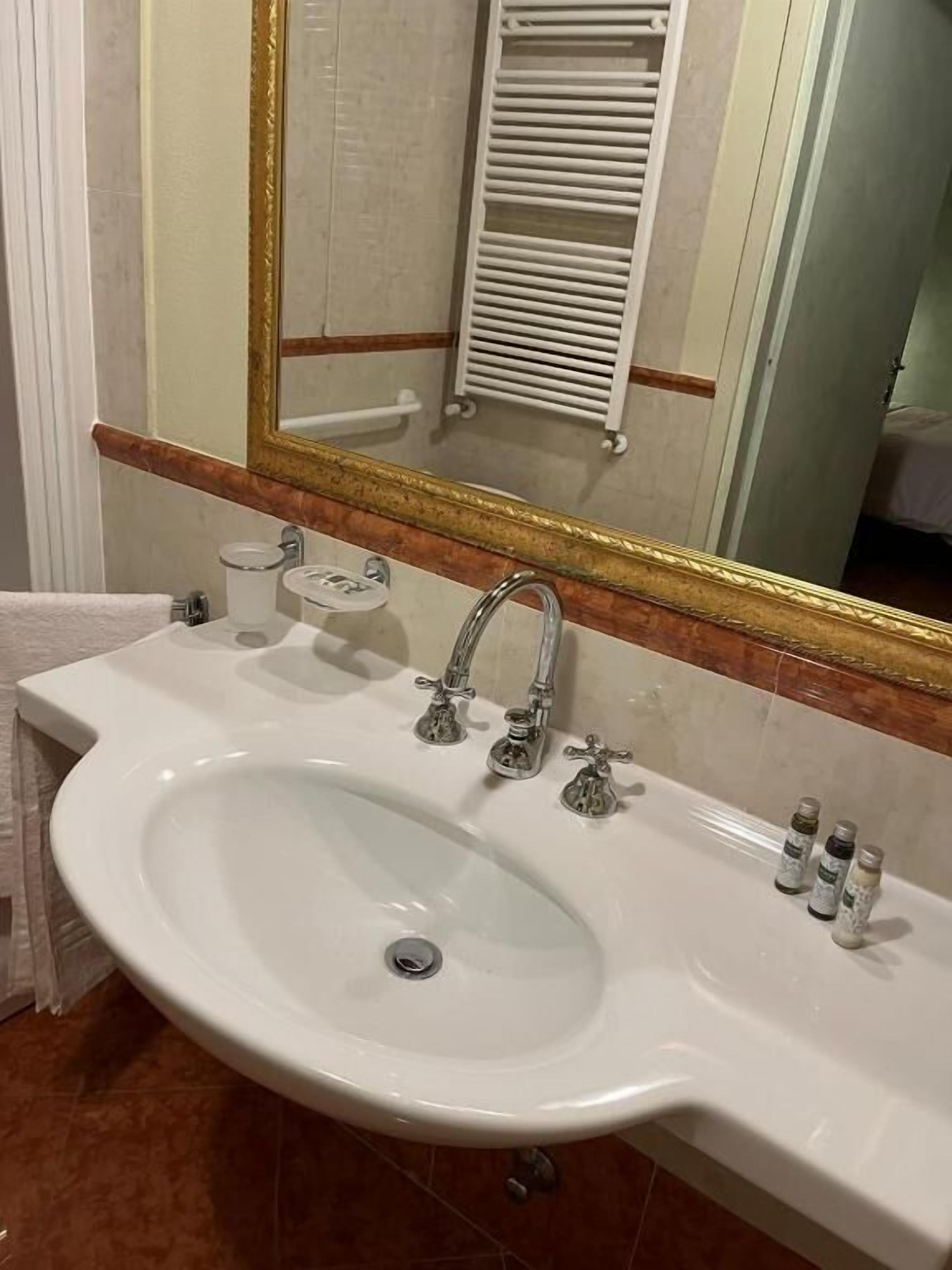 Een badkamer met een wit wastafelmeubel, twee kraanwerkstukken, een spiegel met gouden lijst en een handdoekhouder.
