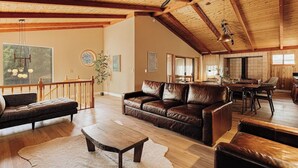 A spacious living room with a wooden ceiling, leather sofas, a wooden coffee table, and a dining area with a large window. - Wildwood Chalet, 5 mins to ski resorts w/game room (Big Bear Lake)