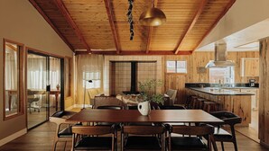 A spacious dining area with a wooden table and chairs, a large pendant light, and a view of the kitchen. - Wildwood Chalet, 5 mins to ski resorts w/game room (Big Bear Lake)