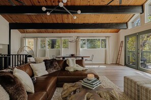 Ein geräumiges Wohnzimmer mit einer braunen Ledersofa, Holzbalkendecke und großen Fenstern.